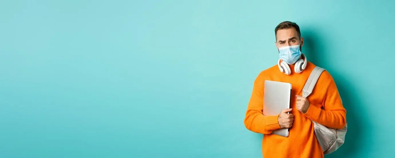Gutaussehender kaukasischer Mann mit Kopfhörern und Rucksack, der einen Laptop hält und eine medizinische Maske trägt, blickt.